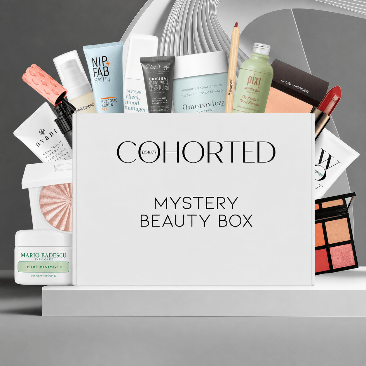 Cohorted Mystery Beauty Box set with a white box in the center and multiple beauty products fanned out behind it, including Nip+Fab, Dr. Brandit, Pixi, Laura Mercier, Mario Badescu, and Estée Lauder, on a gray studio background.